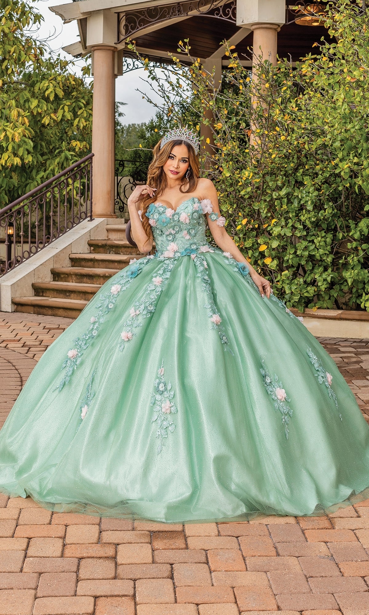 A woman in the Quinceanera Dress 1828 By Dancing Queen, featuring sage green fabric and floral embellishments, stands on a brick pathway before garden stairs, wearing a tiara.